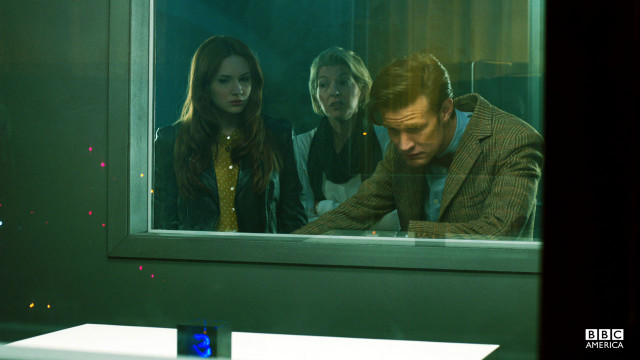The Doctor, Amy, and Kate Stewart stare at a cube.