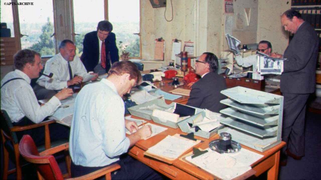 Reporters and journalists at their desks in the newsroom at Alexandra Palace. The windows overlook Alexandra Park. Notice the different color telephones and the fact that everyone is smoking!