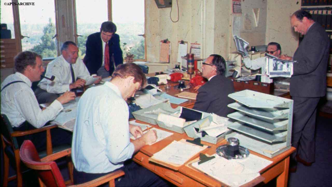 Reporters and journalists at their desks in the newsroom at Alexandra Palace. The windows overlook Alexandra Park. Notice the different color telephones and the fact that everyone is smoking!