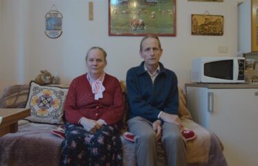 Two people sit on a bed looking straight at the camera.