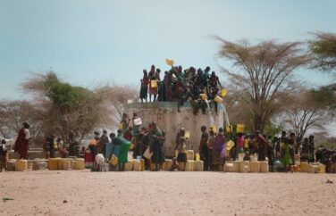 A group of people around and on top of a high well outside on a cloudless day, pour water from the top to those below.