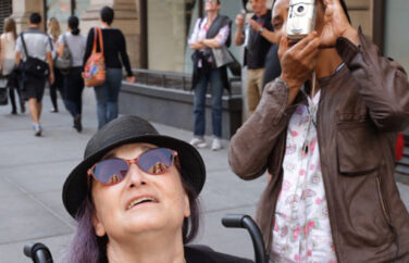 Woman in wheelchair and another person behind her with a camera looking up.