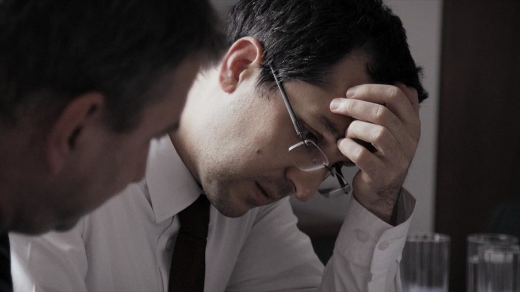 Two men speak, one out of focus, the other in a white dress shirt and glass with his hand on his forehead.