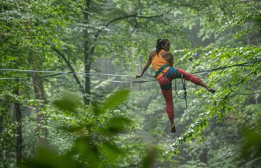 Woman straddles a wire in the middle of a forest.