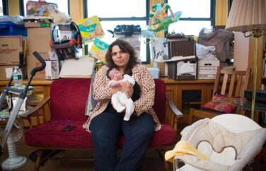 Woman holding a baby sits on a couch surrounded by many boxes.