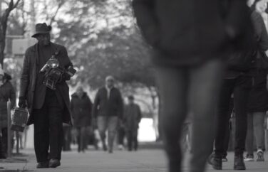 A man walks down a busy sidewalk with a large camera and case.