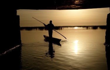 A person in silhouette, upright on a thin boat paddling on the water.