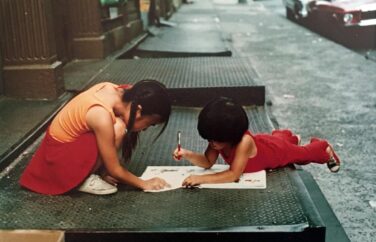 Two children look at a book together outside on a city sidewalk.