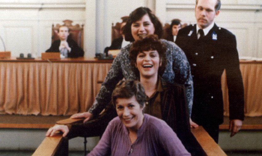 Image is from the film 'A Question of Silence' (1982). Three women walk towards the camera down the center aisle of a courtroom. They are all laughing. An unamused male police officer stands behind them.